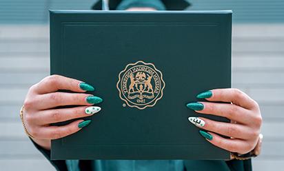 woman holding diploma cover; fingernail polish is green 和 white 和 one nail has a large E
